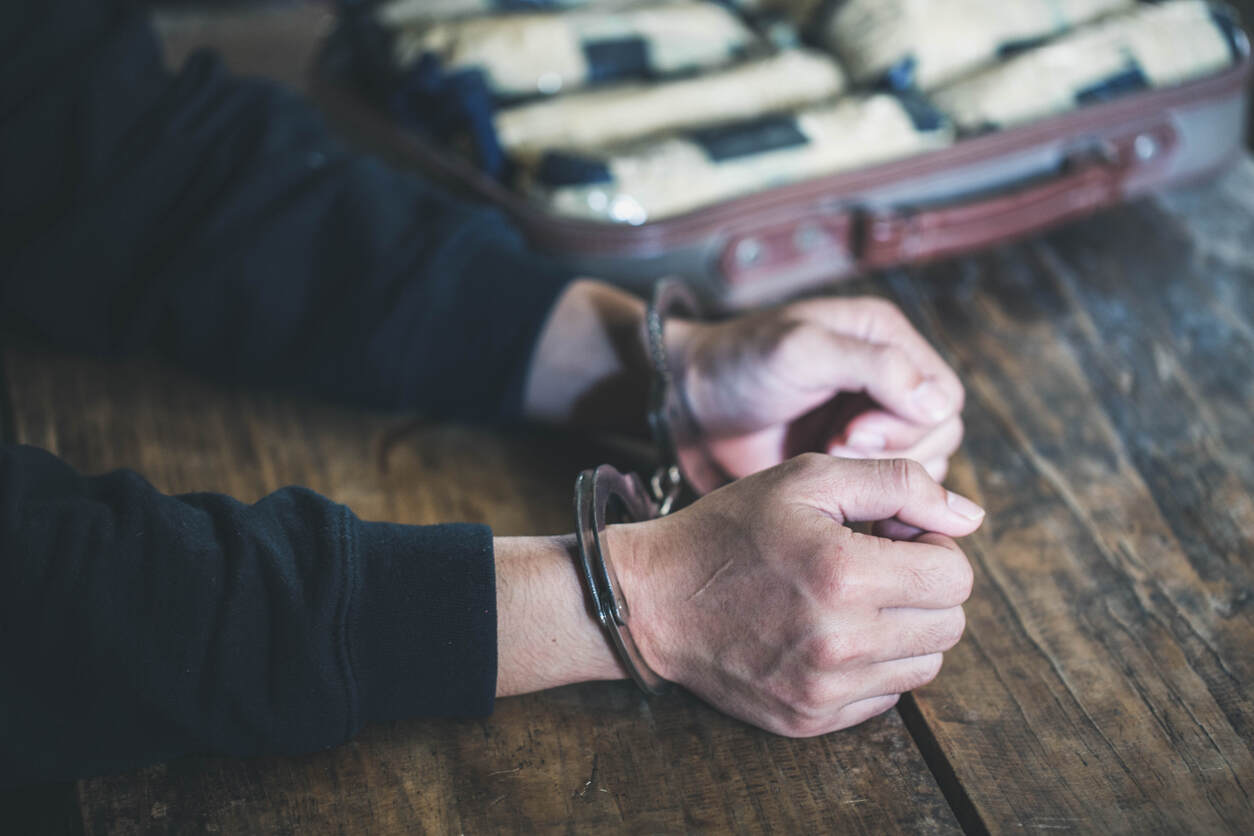 Close-up of handcuffed wrists on a table, next to a suitcase filled with illegal substances, representing a drug trafficking arrest.