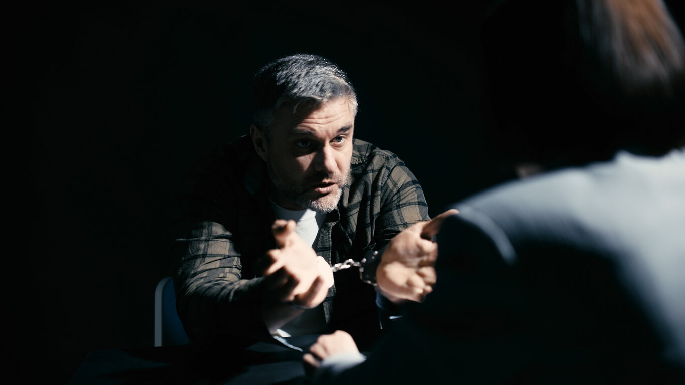 A man in handcuffs speaking to another person across a table in a dimly lit room, symbolizing a legal interrogation or interview related to felony charges.